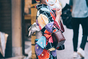 Une femme porte un kimono au motif de fleurs multicolore, elle a un masque de renard devant le visage